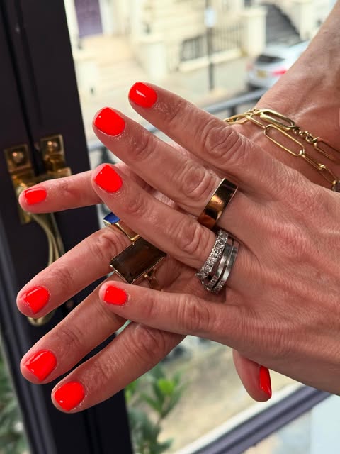 Bright coral-red nails on two hands wearing layered rings and a gold bracelet.