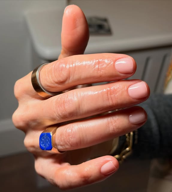 Short, pale pink nails with a glossy finish, photographed with silver and blue-stone rings.